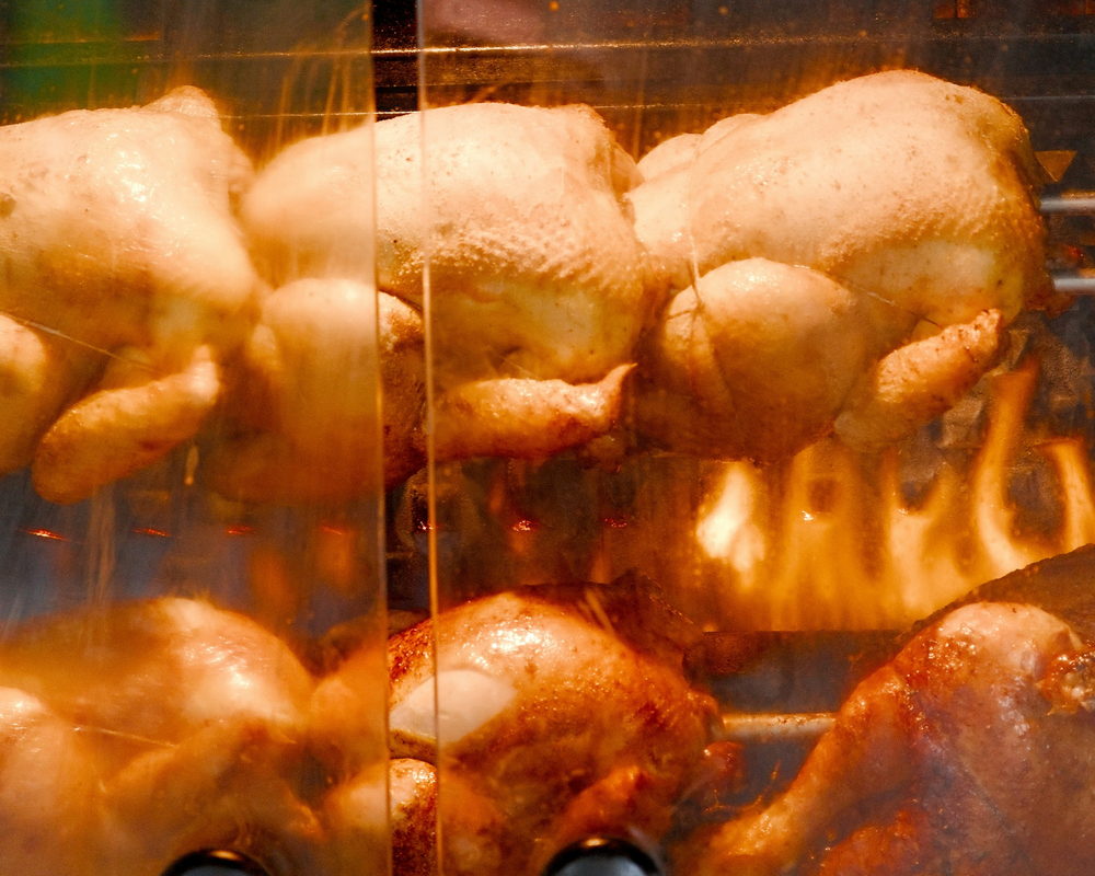 Varios pollos rostizándose en el asador giratorio detrás del cristal, con las llamas encendidas.